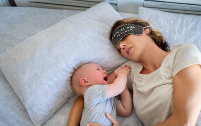 Tired mother with eye band trying to sleep while little toddler crying on bed. Angry baby boy crying and trying to wake mother. Stressed mother trying to take a nap with her child.